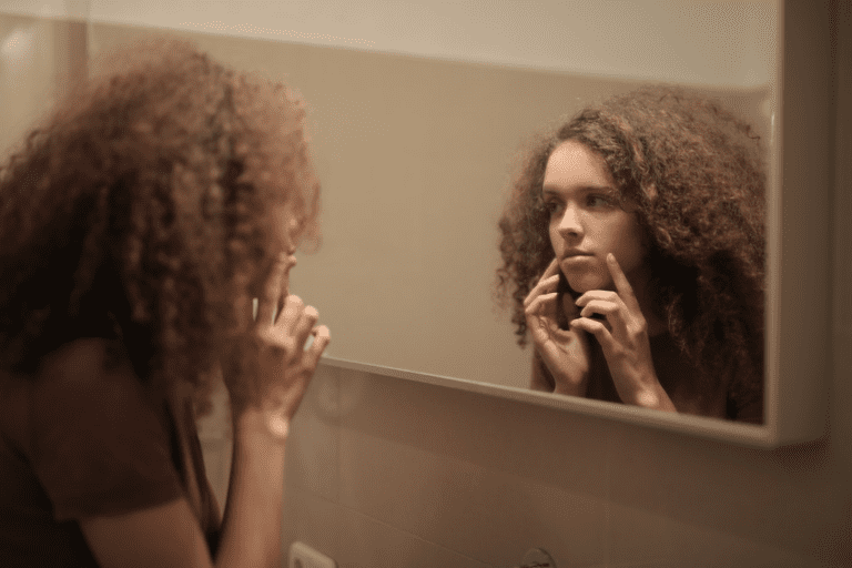 Jovem mulher com cabelo cacheado tocando o rosto enquanto olha no espelho do banheiro.
