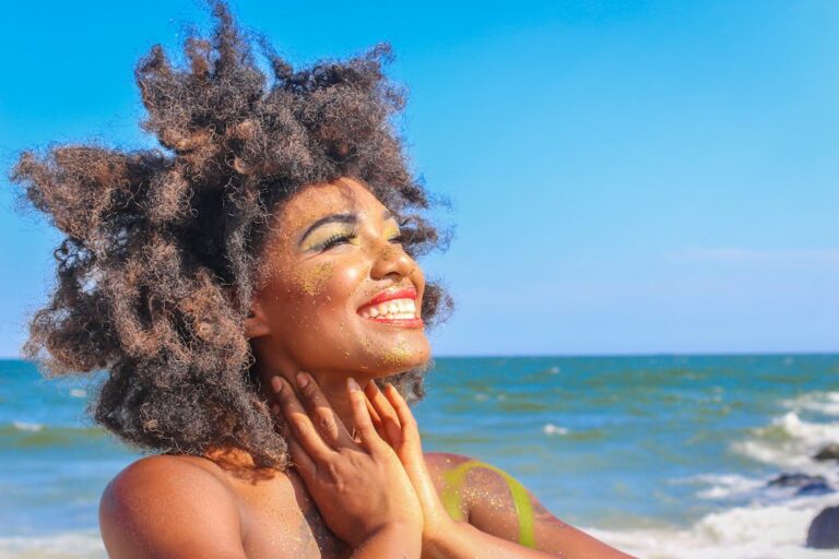Mulher jovem com cabelos cacheados e volumosos sorrindo e segurando seu rosto enquanto está na praia, com o mar ao fundo.